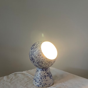 Könnte beinhalten: Eine kugelförmige Tischleuchte mit blau-weißem Sprenkelmuster. Die Leuchte hat eine kreisförmige Öffnung auf der Vorderseite, die ein helles, warmes Licht ausstrahlt. Sie verfügt über einen konischen Fuß und ein weißes Stromkabel an der Rückseite.