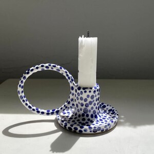 Op de afbeelding: Een keramische kandelaar met een witte basis en een blauw stippenpatroon. Het object heeft een cirkelvormig handvat aan één kant en een centrale cilindrische houder met een witte kaars. Het ontwerp past bij een handgevormde aardewerkstijl.