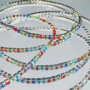 Op de afbeelding: Een set zilverkleurige enkelbandjes met kleurrijke vierkante strasssteentjes. De enkelbandjes zijn gelaagd en hebben verschillende kleuren, waaronder rood, blauw, groen en paars.