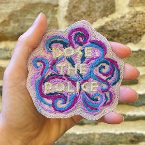 May include: A pink fabric patch with blue and purple embroidery. The patch says "Defund the Police" in gold thread.