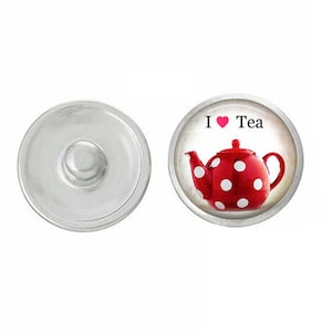 May include: A silver-colored snap button next to a round charm. The charm features a red teapot with white polka dots and the words "I ❤️ Tea". The teapot has a curved spout and handle. The background is white.