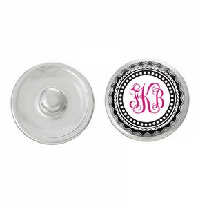 May include: A white snap button with a black and white decorative border and a pink monogram "KB" in the center.