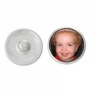 May include: A silver snap button with a photo of a young child smiling. The photo is in a round, silver frame.