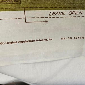 以下が含まれることがあります： テキストと図が描かれた生地のクローズアップ。テキストは「LEAVE OPEN FOR」と「&copy;1983 Original Appalachian Artworks, Inc.」と書かれています。生地は白い背景に緑色のボーダーがあります。