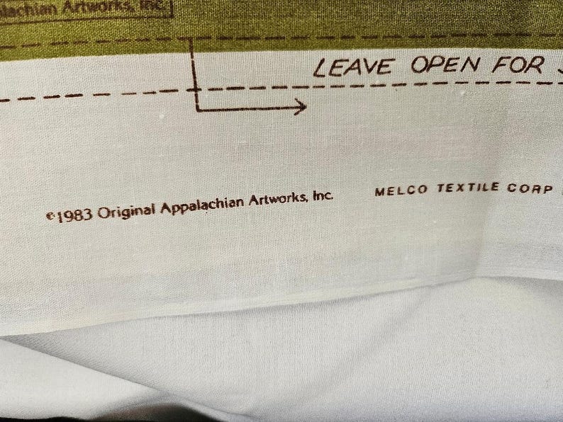 以下が含まれることがあります： テキストと図が描かれた生地のクローズアップ。テキストは「LEAVE OPEN FOR」と「&copy;1983 Original Appalachian Artworks, Inc.」と書かれています。生地は白い背景に緑色のボーダーがあります。