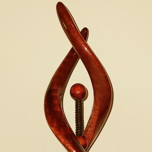 May include: A wooden sculpture with a rich, reddish-brown finish. The abstract design features intertwined, flowing shapes with a central, dark-colored, threaded rod and two spherical accents. The sculpture is mounted on a round base.