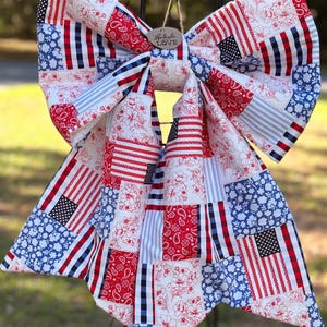 May include: A large, patriotic bow featuring a patchwork design with red, white, and blue patterns. The bow includes American flags, stripes, floral, and paisley prints. A small wooden tag with the words "Handmade LOVE" hangs from the center.