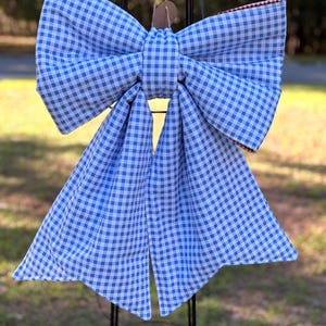 May include: A large, decorative bow made of blue and white gingham fabric. The bow is tied in the centre and has two long, flowing tails. The bow is hanging from a black metal stand, ideal for garden decor.