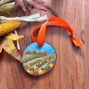 Puede incluir: Un adorno redondo de rodaja de madera con una escena pintada a mano de un campo de calabazas bajo un cielo azul. El adorno tiene un borde de corteza natural y está suspendido por una cinta de satén naranja brillante, con calabazas detalladas.
