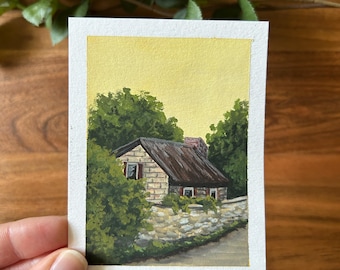 May include: A painting of a stone cottage with a thatched roof, surrounded by green trees and a stone wall. The sky is a pale yellow, suggesting a sunset. The painting is signed "3/30 Cottage".