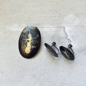 May include: A set of silver earrings and a brooch featuring a black and gold design of a figure with a halo. The earrings are clip-on style.