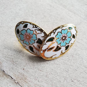 May include: A pair of oval-shaped earrings with a white enamel base and a floral design in shades of blue, green, and pink. The earrings have a gold-tone finish.