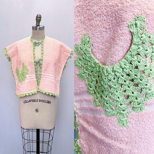 May include: A pink and green crocheted  towel wrap with a 'COLLAPSIBLE SHOULDER' label. The wrap has a green crocheted trim and a green crocheted design on the front.