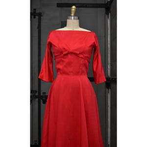 May include: A vibrant red vintage dress with a bateau neckline and three-quarter sleeves. The dress features a fitted bodice with a bow detail and a flared skirt, showcasing a classic silhouette. The dress is displayed on a mannequin.