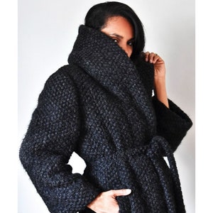May include: A dark gray, textured coat with a wide collar and a matching belt. The coat has long sleeves and pockets. The person in the image is wearing the coat and has their hand in one of the pockets.