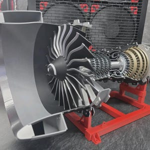 May include: A detailed scale model of a jet engine turbine featuring a large grey intake cowl, a multi-bladed fan, and a complex internal compressor assembly with metallic silver and gold-toned components, all mounted on a bright red plastic stand.