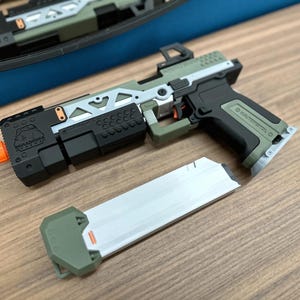 Op de afbeelding: Een speelgoedpistool in olijfgroen en zwart met een zilverkleurig magazijn. Het frame heeft een geometrisch uitgesneden ontwerp, een kleine oranje punt op de loop en een logo met de tekst Paradinha Gear. De grip is gemarkeerd met Maltspistol.