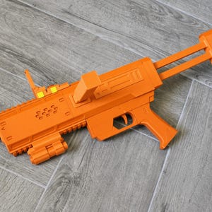 May include: A bright orange toy rifle featuring a top-mounted magazine, a rear stock, a trigger, and a textured barrel. The toy includes small yellow accents on the top sight and a rail system along the bottom of the barrel assembly.