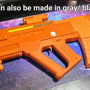 May include: A bright orange toy rifle featuring a tactical rail system, a box magazine, and a small white skull emblem on the side. The text "Can also be made in gray/ black." is printed above the rifle, which includes a yellow accent strip.