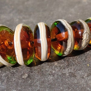 May include: A strand of seven glass beads with a white and brown marbled pattern. The beads are strung on a black cord and have a green and brown iridescent sheen.