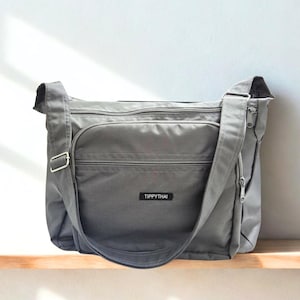 May include: A gray messenger bag with a long adjustable strap and multiple zippered compartments. The bag has a rectangular shape with a front pocket and a small label that reads "TIPPYTHAI". The bag is made of a durable, water-resistant material.