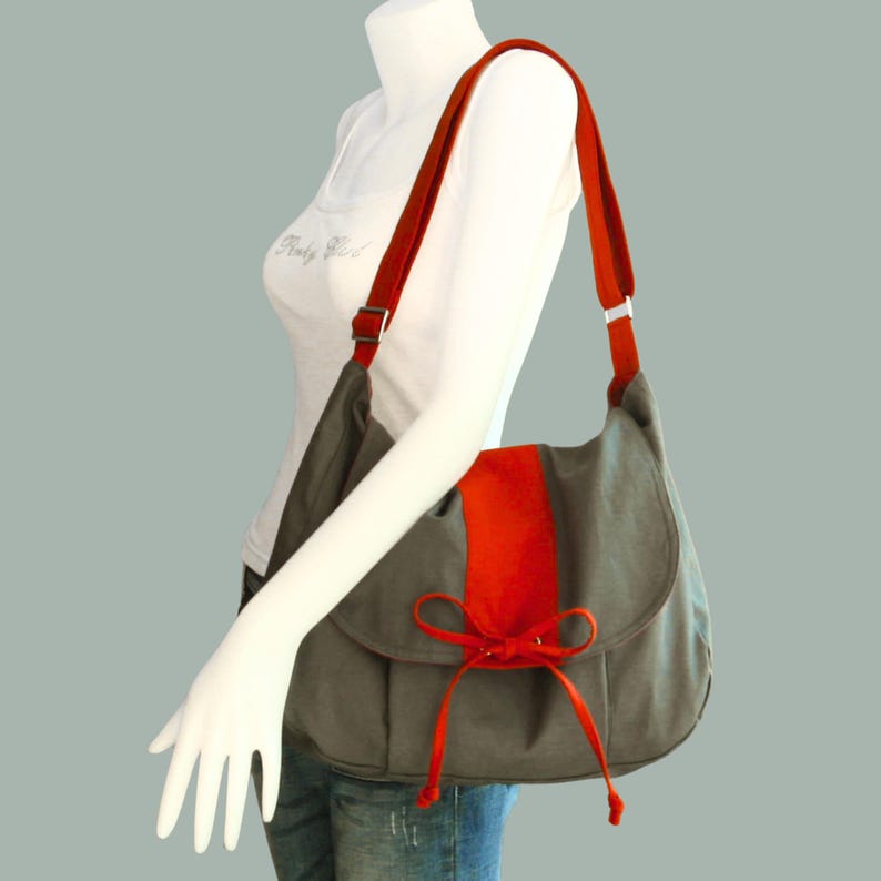 May include: A gray canvas shoulder bag with a red accent and a bow. The bag has a long strap and a flap closure.