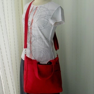 May include: A red canvas tote bag with a long strap. The bag has a zipper closure and two pockets on the front. The bag is being worn by a person wearing a white shirt with a black and red pattern.