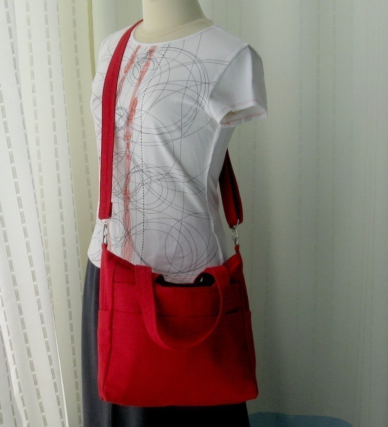 May include: A red canvas tote bag with a long strap. The bag has a zipper closure and two pockets on the front. The bag is being worn by a person wearing a white shirt with a black and red pattern.