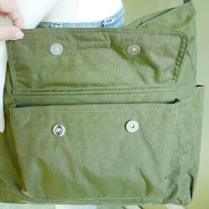 K&ouml;nnte beinhalten: Eine gr&uuml;ne Canvas-Tragetasche mit Klappenverschluss und zwei gro&szlig;en Taschen. Die Tasche hat silberne Metallschnallen und einen Rei&szlig;verschluss.