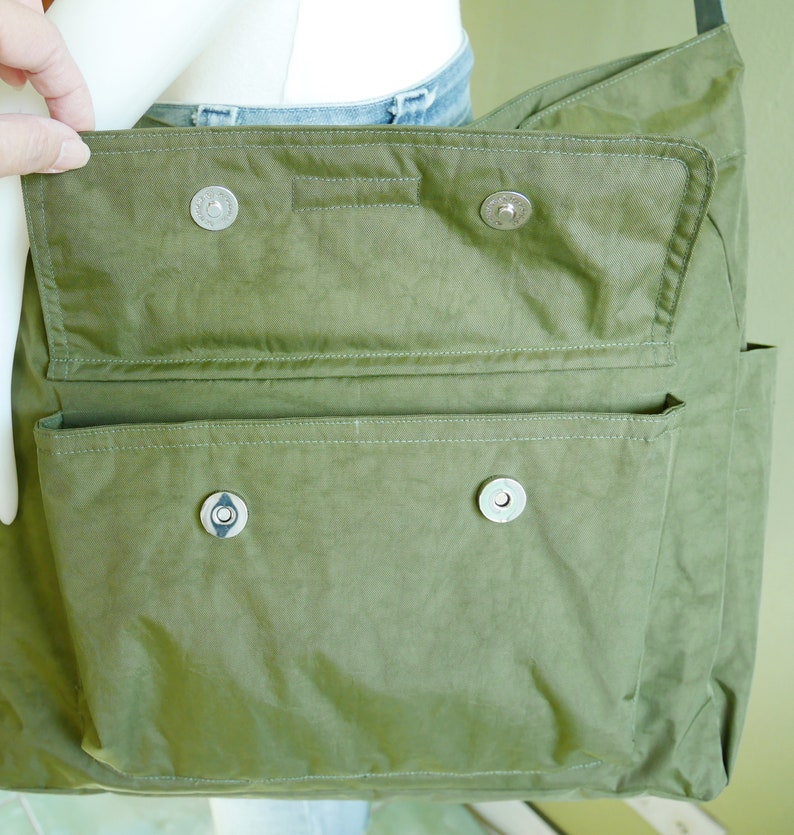 K&ouml;nnte beinhalten: Eine gr&uuml;ne Canvas-Tragetasche mit Klappenverschluss und zwei gro&szlig;en Taschen. Die Tasche hat silberne Metallschnallen und einen Rei&szlig;verschluss.