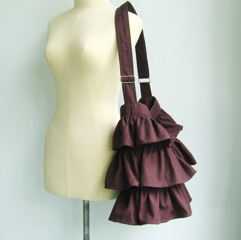 May include: A brown ruffled tote bag with a long strap. The bag is made of fabric and has a simple design. The bag is displayed on a mannequin against a white background.