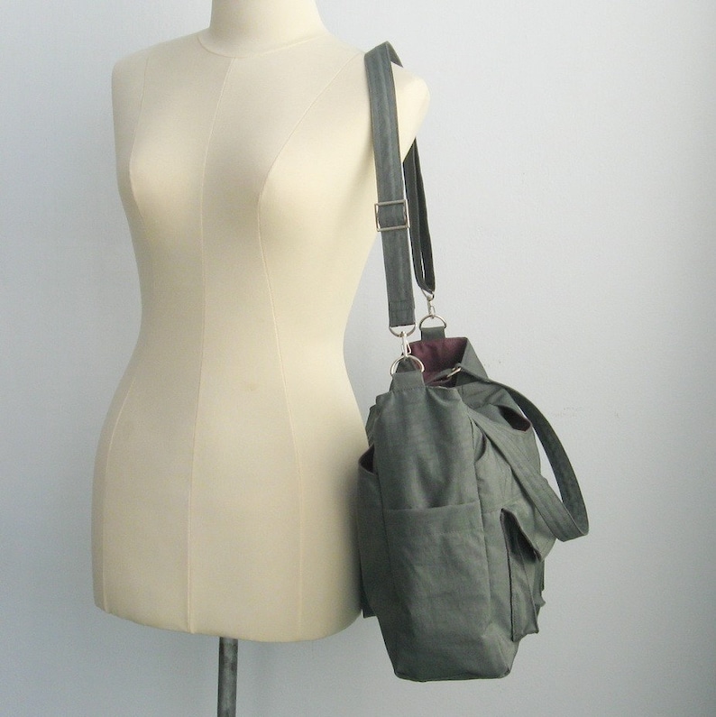 May include: A green canvas shoulder bag with multiple pockets and a long adjustable strap. The bag is displayed on a mannequin and features a silver buckle and hardware. The interior lining is a deep burgundy color.