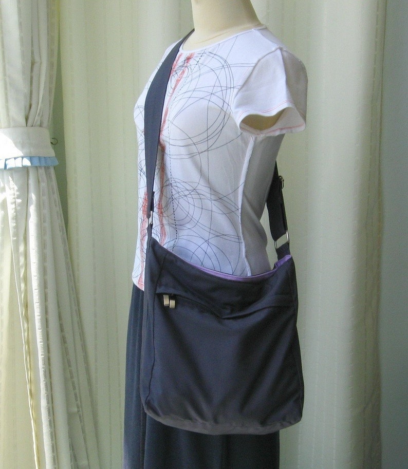 May include: A dark blue crossbody bag with a purple lining and a silver zipper. The bag is worn over a white shirt with a black and red pattern.