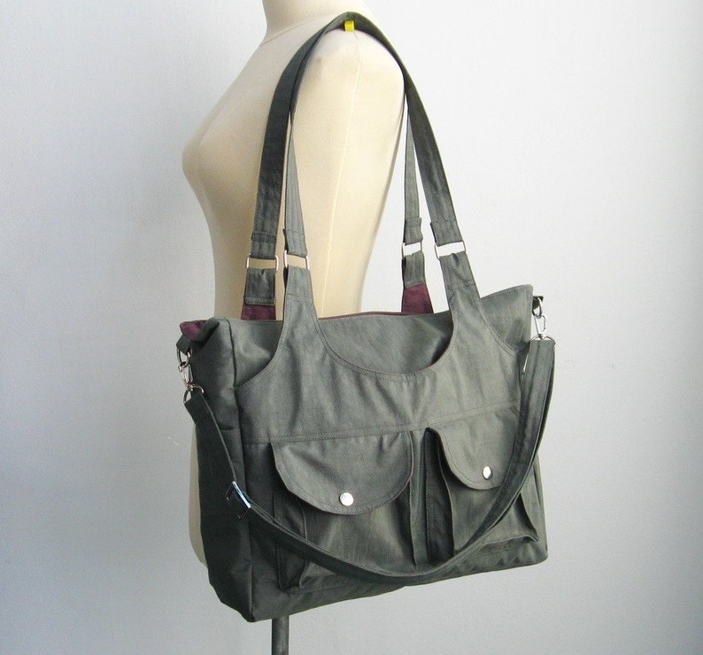 May include: Olive green tote bag with two front pockets and a shoulder strap. The bag has two top handles and a burgundy interior lining. The bag is displayed on a mannequin.