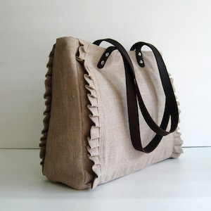 K&ouml;nnte beinhalten: Beige Tragetasche mit braunen Ledergriffen. Die Tasche hat einen ger&uuml;schten Rand an den Seiten. Die Griffe sind mit silbernen Nieten befestigt. Die Tasche ist aus einem strukturierten Stoff gefertigt.