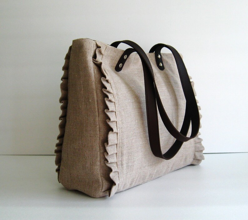 K&ouml;nnte beinhalten: Beige Tragetasche mit braunen Ledergriffen. Die Tasche hat einen ger&uuml;schten Rand an den Seiten. Die Griffe sind mit silbernen Nieten befestigt. Die Tasche ist aus einem strukturierten Stoff gefertigt.