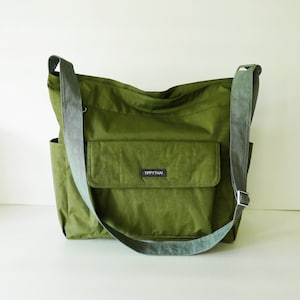 K&ouml;nnte beinhalten: Eine gr&uuml;ne Canvas-Schultertasche mit einer gro&szlig;en Fronttasche und einer kleineren Tasche an der Seite. Die Tasche hat ein schwarzes Etikett mit der Aufschrift "Tippythai".