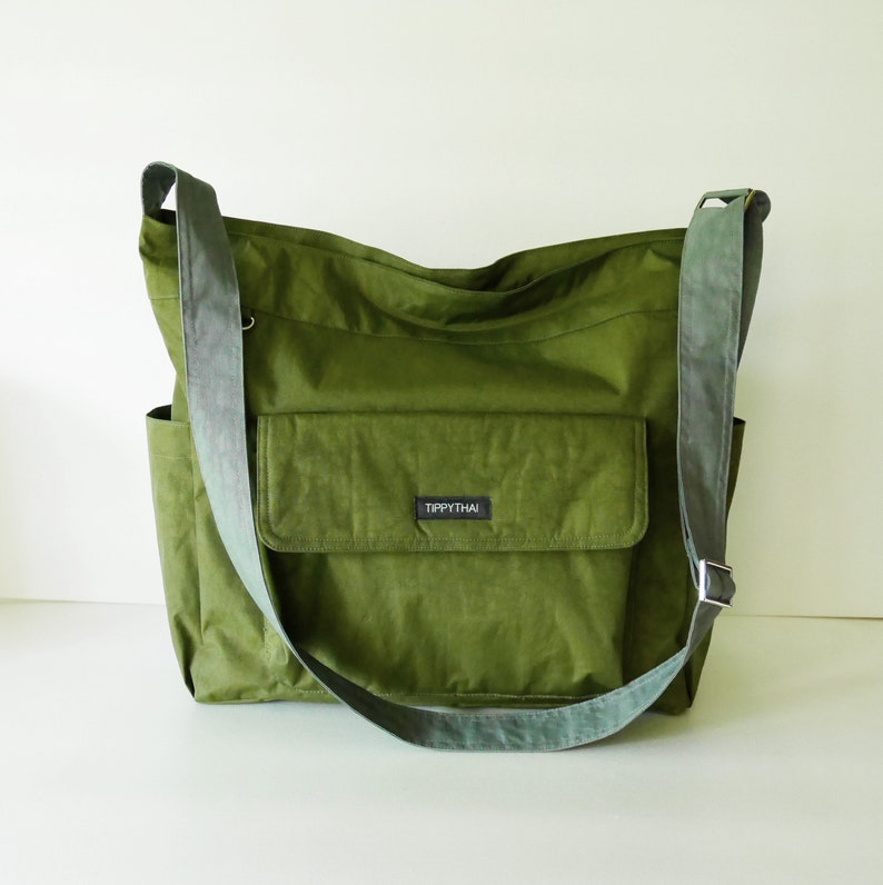 K&ouml;nnte beinhalten: Eine gr&uuml;ne Canvas-Schultertasche mit einer gro&szlig;en Fronttasche und einer kleineren Tasche an der Seite. Die Tasche hat ein schwarzes Etikett mit der Aufschrift "Tippythai".
