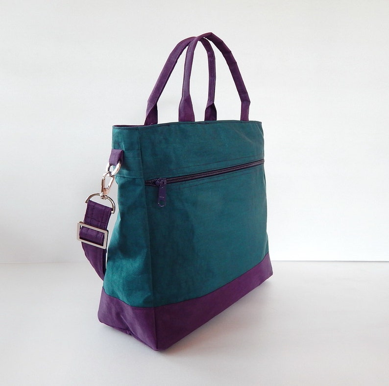 May include: A teal green tote bag with a purple bottom and a purple strap. The bag has a zippered pocket on the front.