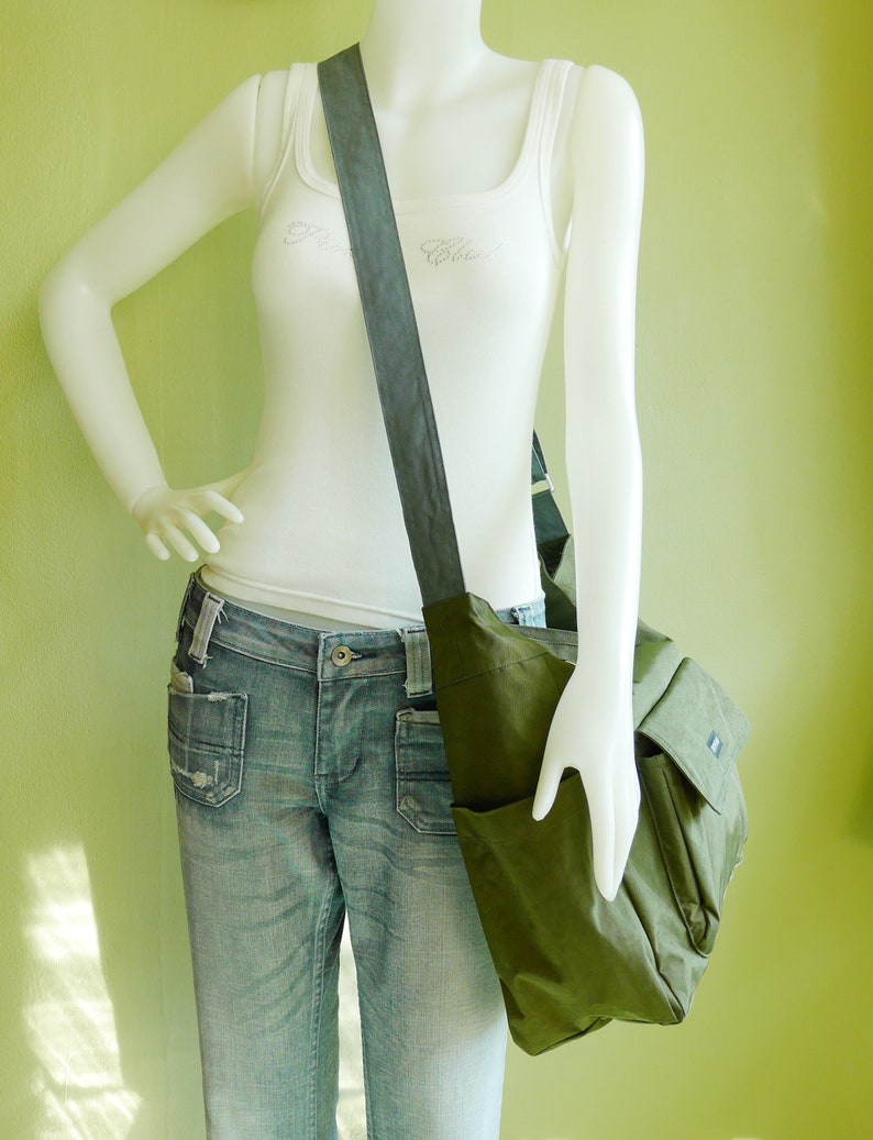 K&ouml;nnte beinhalten: Eine gr&uuml;ne Canvas-Messenger-Tasche mit einem langen, verstellbaren Trageriemen. Die Tasche hat einen Klappverschluss und eine Fronttasche. Die Tasche wird von einer Schaufensterpuppe getragen.