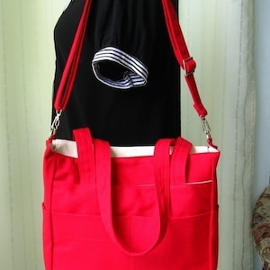 May include: A red canvas tote bag with a white lining and two top handles. The bag has a long adjustable strap and multiple pockets.