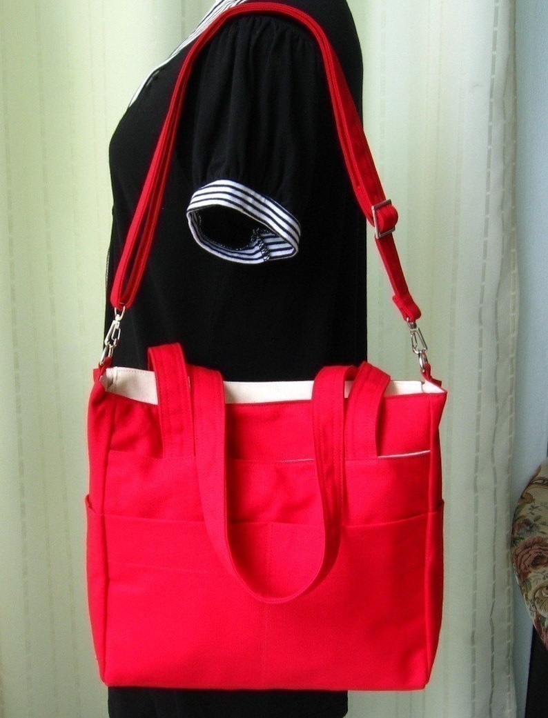 May include: A red canvas tote bag with a white lining and two top handles. The bag has a long adjustable strap and multiple pockets.