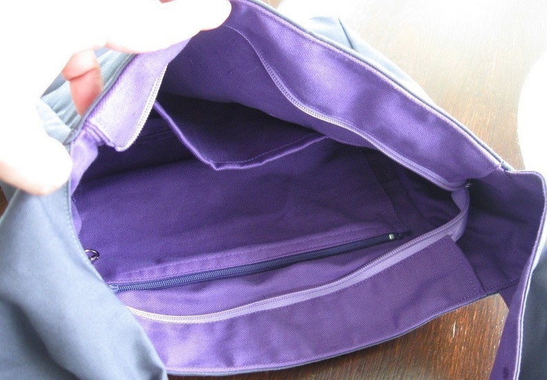 May include: A gray canvas tote bag with a purple lining. The bag has a zippered pocket and two open pockets inside.