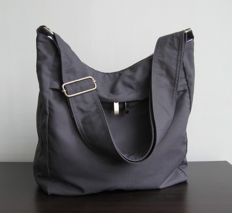 May include: A dark gray canvas tote bag with a long adjustable strap and a silver buckle. The bag has a flap with a small pocket on the front.