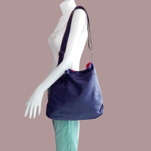 May include: A dark blue canvas tote bag with a red lining and a long adjustable strap. The bag is worn over the shoulder of a mannequin.