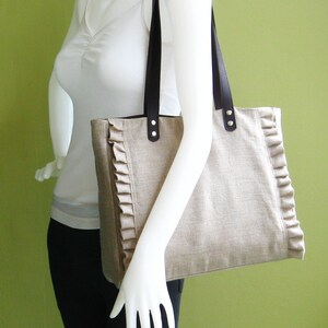 K&ouml;nnte beinhalten: Beige Tragetasche mit dunkelbraunen Ledergriffen und dekorativen R&uuml;schen an den Kanten. Die Tasche wird von einer Schaufensterpuppe gehalten. Die Tasche hat eine rechteckige Form und ist aus einem strukturierten Stoff gefertigt.
