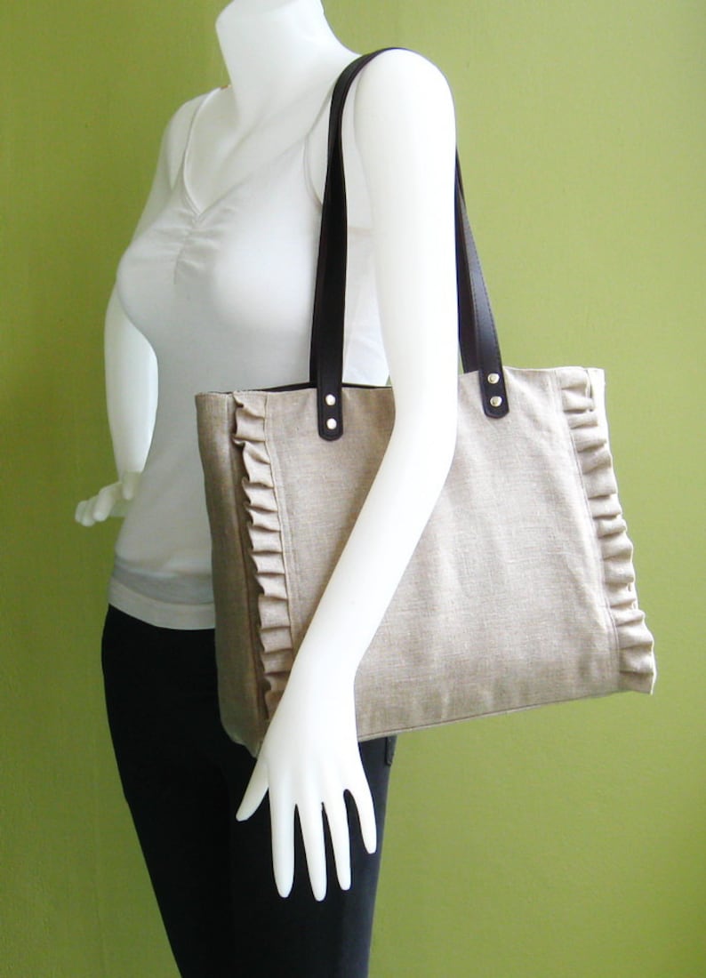 K&ouml;nnte beinhalten: Beige Tragetasche mit dunkelbraunen Ledergriffen und dekorativen R&uuml;schen an den Kanten. Die Tasche wird von einer Schaufensterpuppe gehalten. Die Tasche hat eine rechteckige Form und ist aus einem strukturierten Stoff gefertigt.