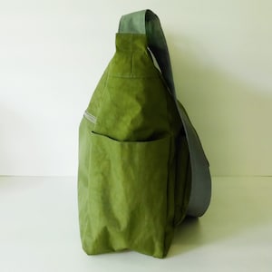 K&ouml;nnte beinhalten: Eine gr&uuml;ne Canvas-Schultertasche mit einem grauen Riemen. Die Tasche hat eine gro&szlig;e Tasche auf der Vorderseite und eine Rei&szlig;verschlusstasche an der Seite.