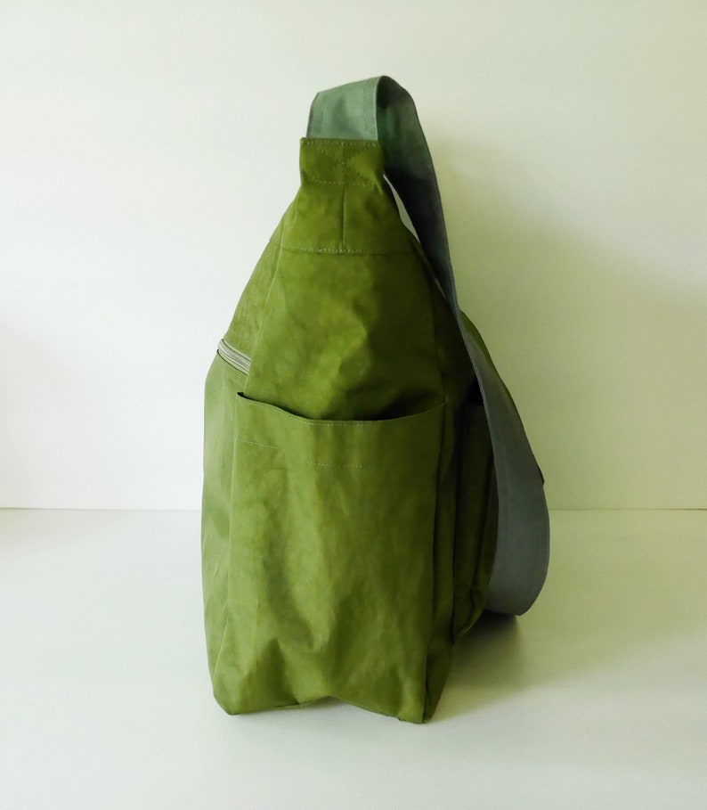 K&ouml;nnte beinhalten: Eine gr&uuml;ne Canvas-Schultertasche mit einem grauen Riemen. Die Tasche hat eine gro&szlig;e Tasche auf der Vorderseite und eine Rei&szlig;verschlusstasche an der Seite.