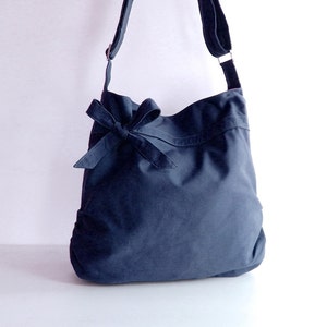 May include: A dark blue canvas shoulder bag with a large bow on the front. The bag has a long adjustable strap.